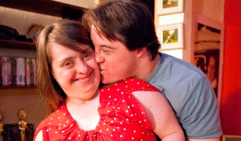 Um jovem casal com Síndrome de Down. Rapaz beijando o rosto da namorada e a abraçando por trás