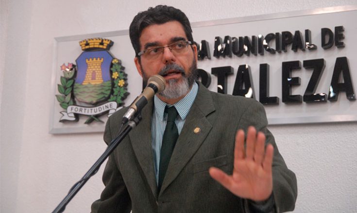 Homem de barba grisalha, óculos e usando paletó e gravata discursa ao microfone à frente de uma placa dizendo Câmara Municipal de Fortaleza