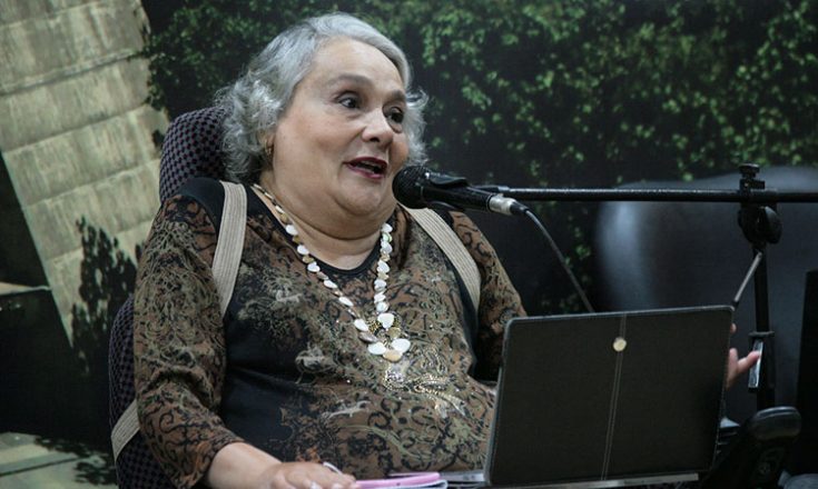 Mulher de meia idade, cabelos brancos, falando ao microfone