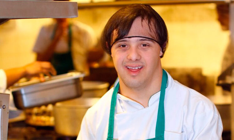 Rapaz com síndrome de down, vestido com uniforme de cozinha e uma touca nos cabelos, sorrindo