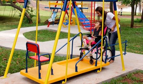 Parque de diversão em que uma garotinha, em uma cadeira de rodas, brinca em um balanço amarelo adaptado, empurrada por uma mulher