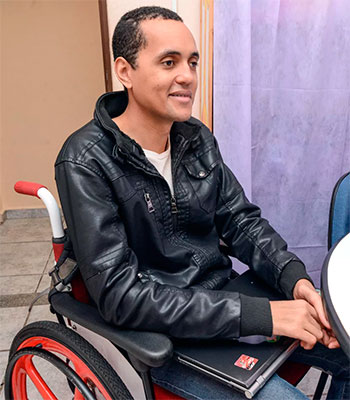 Homem jovem, sentado em uma cadeira de rodas, vestindo uma jaqueta jeans preta e segurando um laptop nas pernas