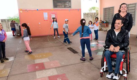 Pátio de uma escola com uma mulher empurrando uma cadeira de rodas com um homem sentado e, ao fundo, crianças brincam de pular corda, conversam e bebem água no bebedouro