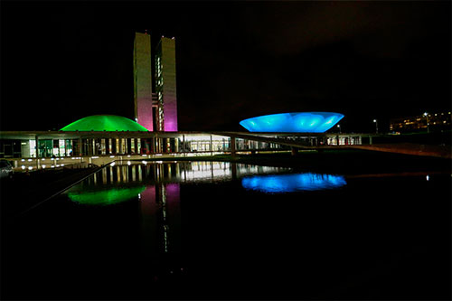 Foto escura das cúpulas do Congresso Nacional iluminadas de verde, azul, rosa e roxo