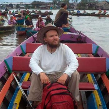 Voto quadrada de um homem em um barco, com uma mochila vermelha a sua frente. Ao fundo, vários outros barcos e pessoas navegando