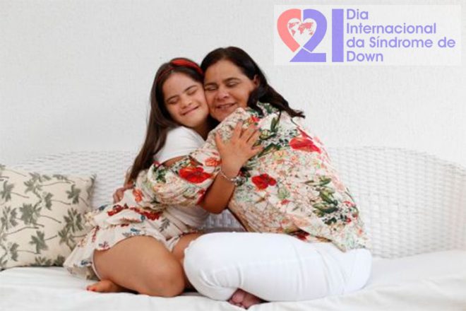 Uma mulher abraçada a sua filha adolescente e com Síndrome de Down, ambas sentadas em um sofá. No alto, à direita, a logomarca do Dia Internacional da Síndrome de Down.