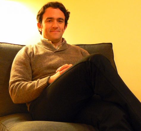 Foto quadrada de um homem sentado em um sofá, com a perna direita cruzada sobre a esquerda, as mãos cruzadas sobre a coxa direita e, ao fundo, uma parede amarela e o reflexo de uma luz no canto superior esquerdo
