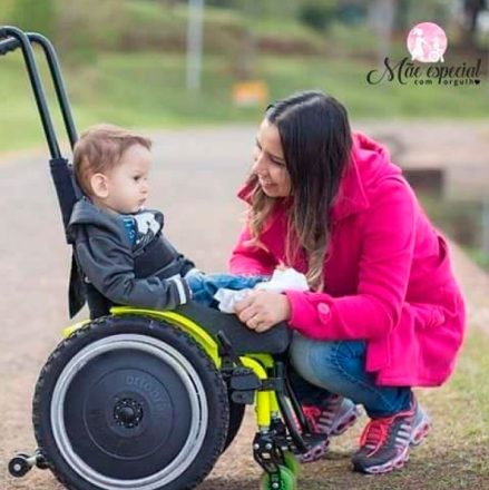Mãe ajoelhada e sorrindo para seu filho em uma cadeira de rodas, de frente para ele. No alto, à direita, a logo do blog Mães Especiais com Orgulho