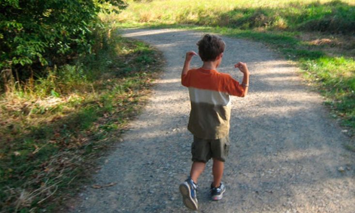 Foto horizontal de um menino autista de cerca de dez anos, de costas, vestindo uma bermuda escura, tênis azul e uma blusa tricolor em laranja, branco e marrom, caminhando por uma estrada cercada por vegetação dos lados, e realizando movimentos repetitivos com os braços.