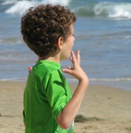 Foto quadrada, da cintura para cima, de um menininho autista, olhando para o mar. Ele veste uma camiseta verde e realiza o chamado flapping, movimento repetitivo com os braços.