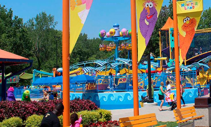 Imagem do parque de diversões Sesame Place, com postes laranjas e flâmulas de animais, em primeiro plano, e brinquedos e pessoas passeando, ao fundo.