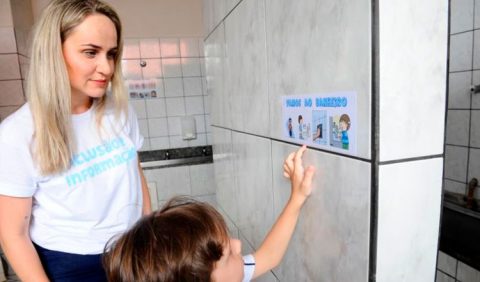 Foto de uma mãe loira, vestindo uma camiseta branca com os dizeres INCLUSÃO E INFORMAÇÃO, em azul, junto com seu filho. O garoto aponta para um adesivo na parede, informando que ali é a entrada do banheiro da escola