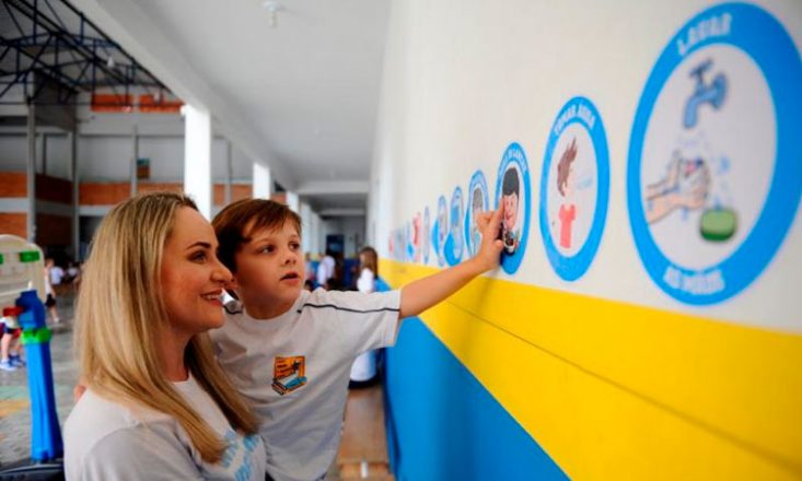 Mãe segura o filho autista no colo e ele aponta para vários adesivos pregados na parede da escola, indicando atividades rotineiras do garoto.