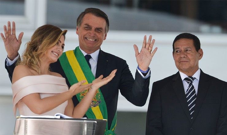 Três pessoas de frente para a foto. À esquerda, uma mulher de vestido claro sorri e aponta as mãos para o homem ao centro, de paletó e a faixa presidencial ao peito e um segundo homem, à direita, também de paletó.