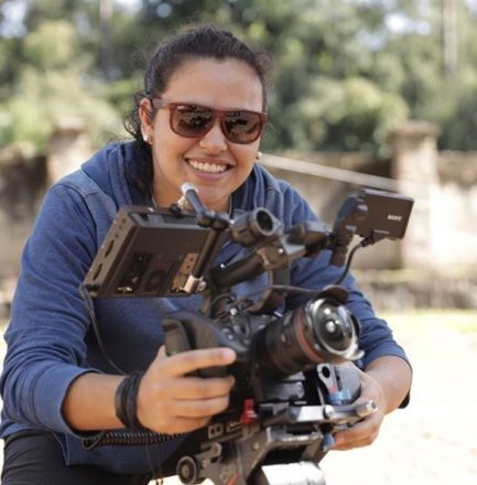 Mulher de óculos escuros, sorrindo, segura uma câmera de filmagem, de frente para a foto.