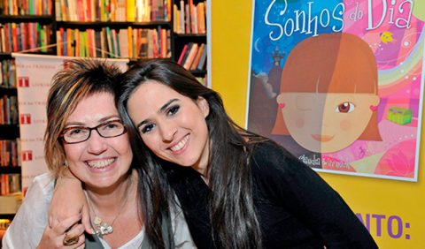 Mãe e filha se abraçam e sorriem em uma livraria. Ao fundo, uma estante de livros e um cartaz. À direita, a capa do livro Sonhos do Dia, em formato ampliado.