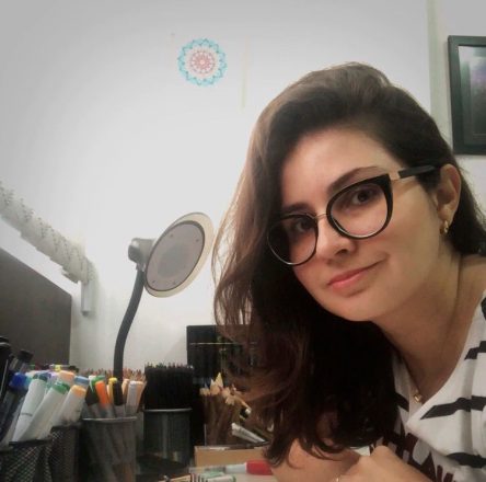 Foto quadrada de uma jovem sorrindo, de óculos, em um escritório, cercada de lápis coloridos organizados em pequenos potinhos.