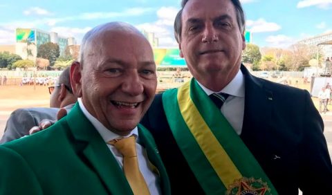 Dois homens, lado a lado, o empresário Luciana Hang, à esquerda, de paletó verde, blusa branca e gravata amarela e Jair Bolsonaro à direita, de paletó escuro e a faixa presidencial no peito.