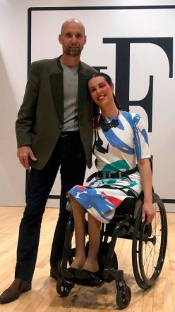 Foto vertical de um homem, de pé, careca, de blazer marrom, abraçado a uma mulher jovem e sorridente, em uma cadeira de rodas, usando um vestido colorido em branco, azul, verde e vermelho.