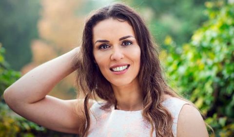 Imagem em foco de uma mulher jovem, bonita, sorrindo, ajeitando os cabelos esvoaçantes. Ao fundo, muitas plantas.